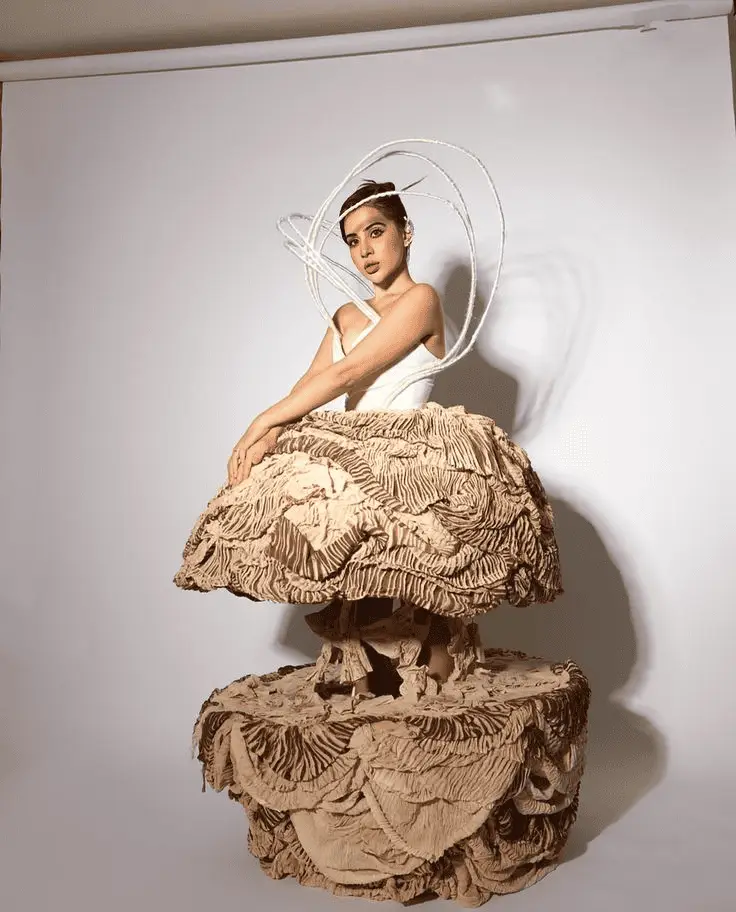 Urfi wearing a sculptural layered beige ruffled dress with dramatic circular headpiece, posed against a plain white studio backdrop, creating an artistic high-fashion editorial look.