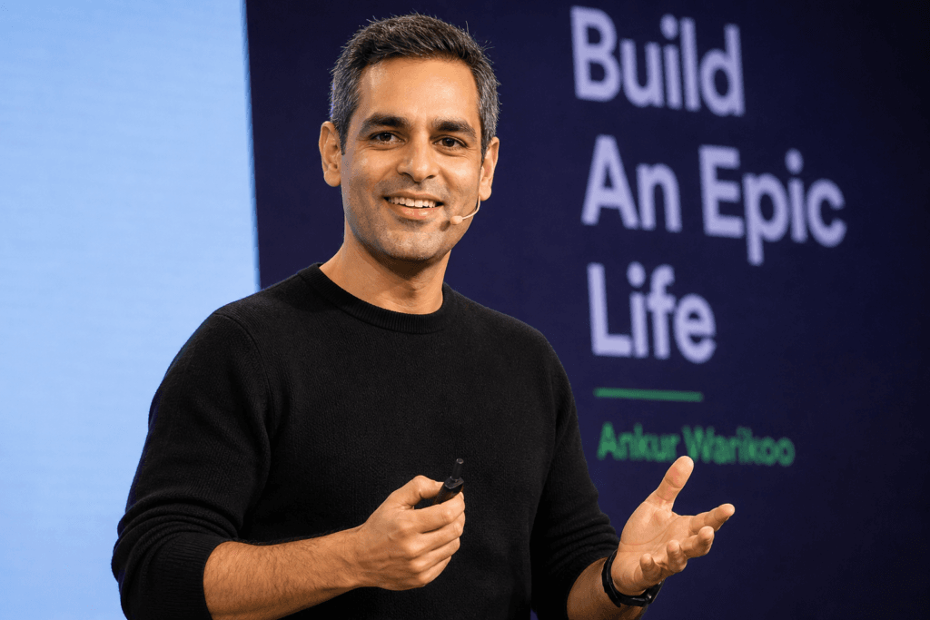 A professional image of Ankur Warikoo speaking on stage, smiling confidently while holding a presentation remote, with a blurred background displaying the words “Build An Epic Life,” reflecting his role as a motivational speaker and digital educator.